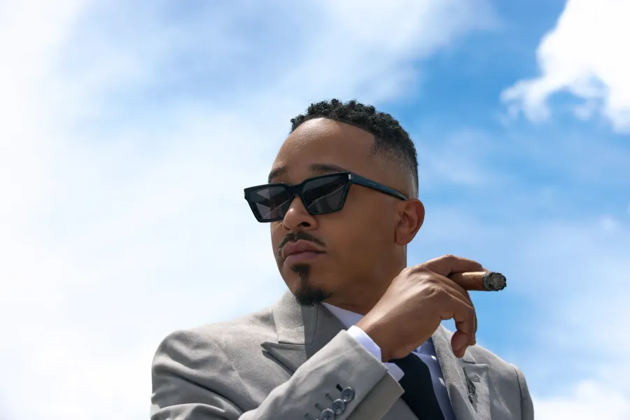 Grey suit and sunglasses, cigar raised against open sky